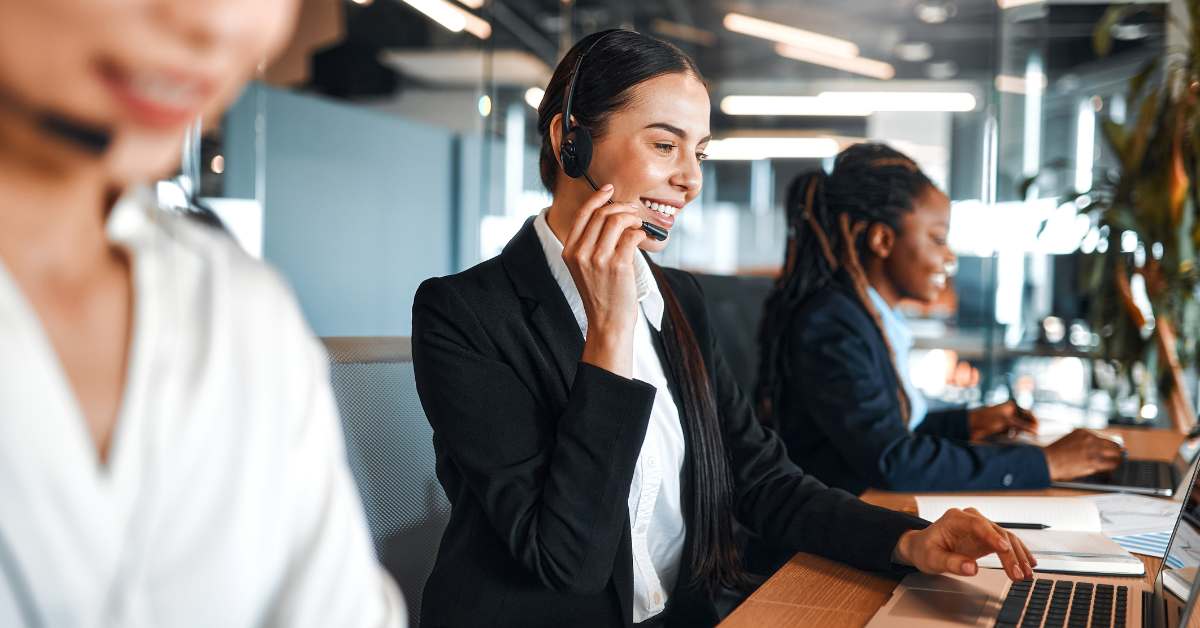Businesswoman wearing a headset smiles while taking a call on a cloud PBX system at her laptop in a modern office.
