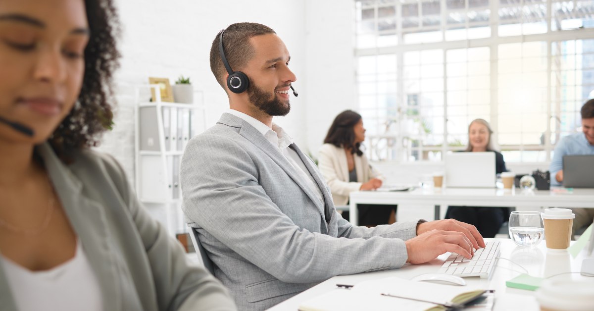 A consultant wearing a grey suit and a headset, seated at his desk, and speaking with a client.