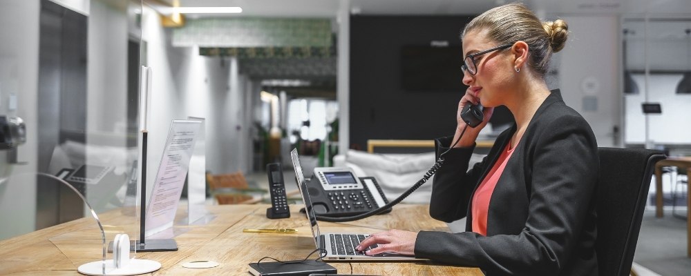 A businesswoman, wearing a suit, seated at her desk, working on her laptop, making a call using her VoIP deskphone and on-premise PBX system.