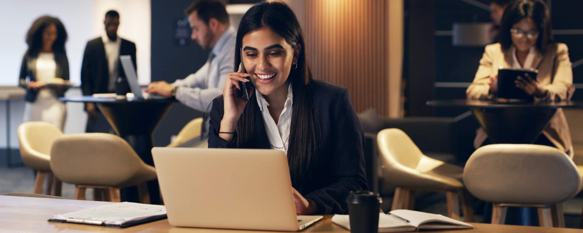 Businesswoman taking a mobile call while working on her laptop in a co-working space, using a cloud-based business phone system.
