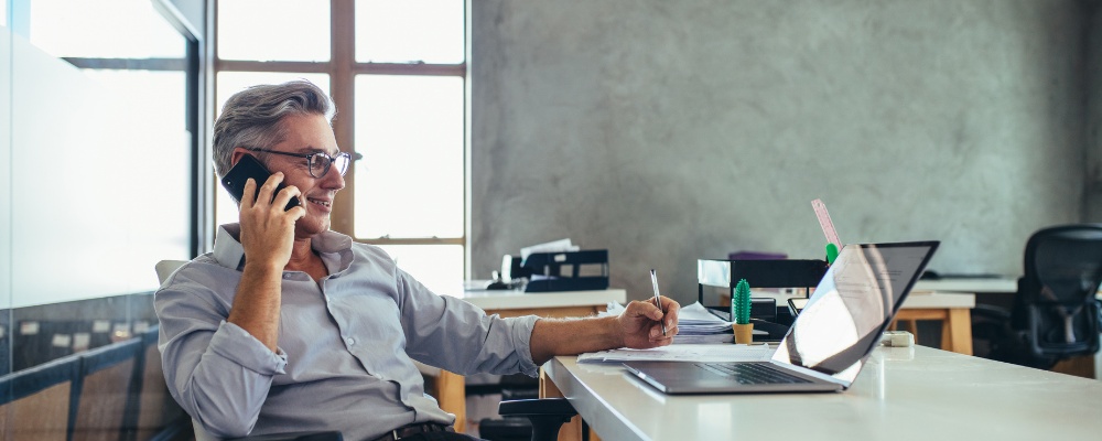 Man in grey shirt seated at his desk making a call with his mobile and a VoIP package