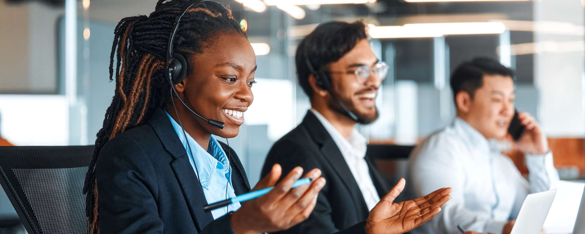 Call centre agent wearing a headset smiles and gestures during a business phone call, with two colleagues also on headsets in the background.